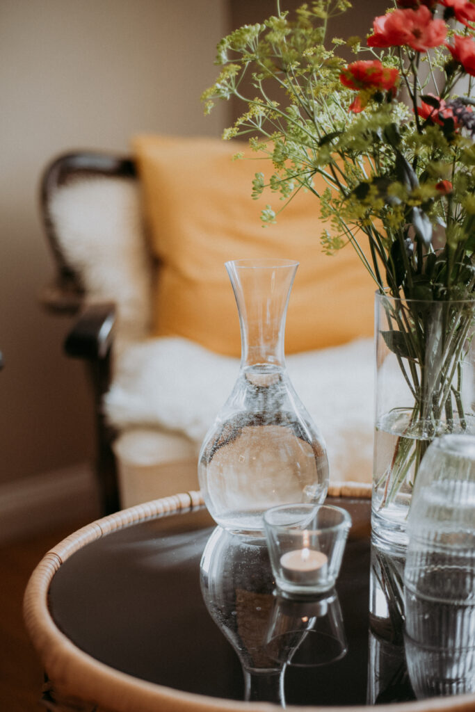 Runder Tisch mit frischem Blumenstrauß aus roten Blümchen und grünem Beiwerk, Glaskaraffe und brennender Teelichtkerze – einladende Dekoration in der Sitzecke.