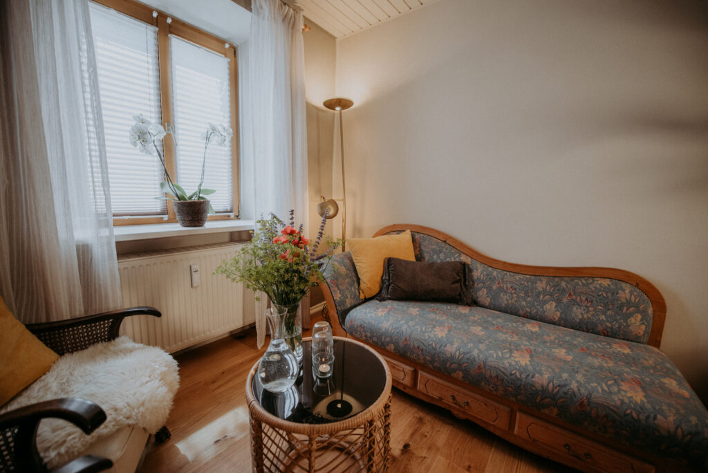 Helle Sitzecke mit floraler Chaiselongue, großem Fenster mit Orchidee, Blumenstrauß auf dem Tisch und warmem Licht – ruhiger und einladender Bereich der Massage- und Tantrapraxis.