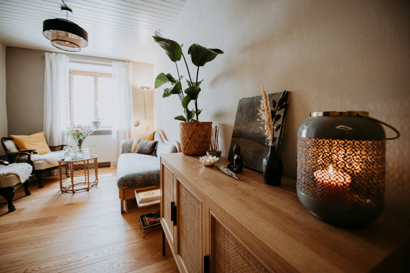 Wohnlicher Wartebereich der Massagepraxis mit hellem Holzsideboard, großer Zimmerpflanze, Kerzenlaterne, Buddha-Figur und gemütlicher Sitzecke mit Tageslicht.